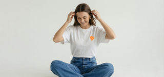 Smiling woman sitting cross-legged wearing white Be Like Children Christian t-shirt with orange heart on front, hands in hair