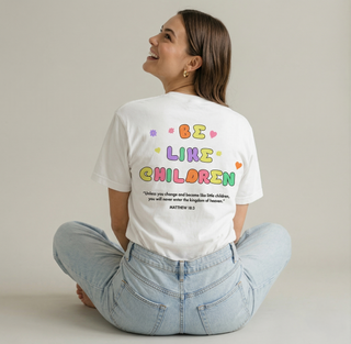 Smiling woman sitting cross-legged seen from behind wearing white Be Like Children Christian t-shirt with colorful back graphic and Matthew 18:3 Bible verse