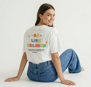 Smiling woman sitting on the ground looking over her shoulder showing the back of white Be Like Children Christian t-shirt with colorful graphic and Matthew 18:3 verse