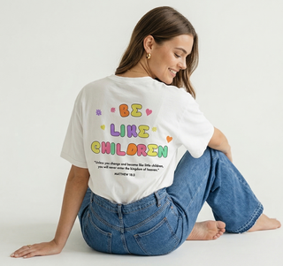 Woman sitting with one knee up showing the back of white Be Like Children Christian t-shirt with colorful graphic print and Matthew 18:3 Bible verse