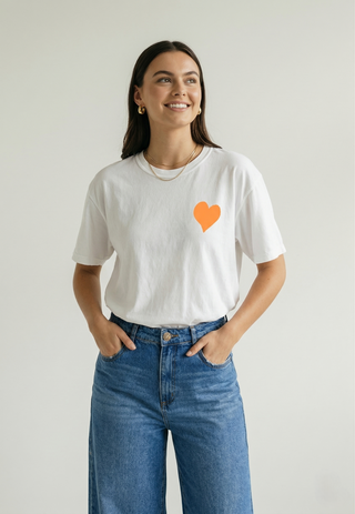 Smiling woman standing with hands in pockets wearing white Be Like Children Christian t-shirt with orange heart graphic on front, full-body front view