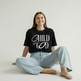 Happy woman sitting in oversized black "Child of God" Christian calligraphy tee with wide-leg light jeans, joyful faith lifestyle photo