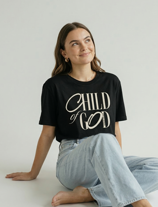 Close-up front view of "Child of God" calligraphy design on black Christian graphic tee, woman looking up with faith and confidence