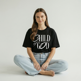 Woman sitting cross-legged in black "Child of God" Christian graphic tee with light wash jeans, women's faith-based clothing