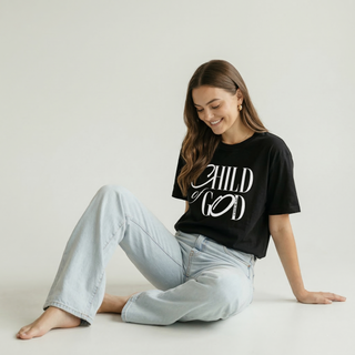 Woman sitting in black "Child of God" Christian calligraphy graphic t-shirt with light wash wide-leg jeans, faith-inspired women's tee