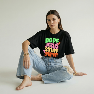 Woman sitting on the floor wearing the Dope Jesus Tee in black with colorful "Dope Jesus Stuff Everyday" graphic print, paired with wide-leg jeans