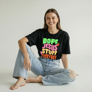 Smiling woman sitting on the floor wearing the Dope Jesus Tee in black with colorful "Dope Jesus Stuff Everyday" graphic print, paired with ripped wide-leg jeans
