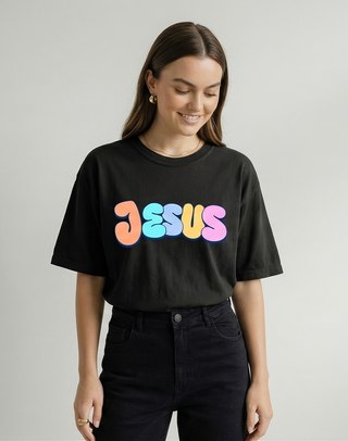 Smiling woman in black oversized "Jesus Loves You" Christian graphic tee with vibrant pastel retro bubble letter JESUS design, black jeans — Faith & Co.