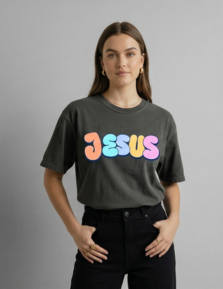 Woman in grey "Jesus Loves You" Christian graphic tee with colorful pastel retro bubble letter JESUS front design, hands in pockets — Faith & Co.