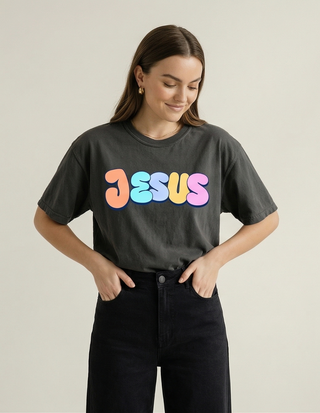 Smiling woman in grey "Jesus Loves You" Christian graphic tee with colorful retro bubble letter JESUS design, hands in jeans pockets — Faith & Co.