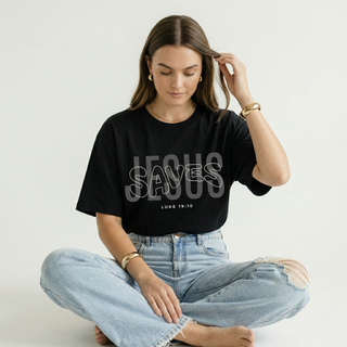 Woman sitting cross-legged in black "Jesus Saves" Luke 19:10 Christian graphic tee with bold layered text design, paired with wide-leg ripped denim