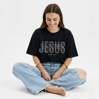 Woman sitting cross-legged on white background in black "Jesus Saves" Luke 19:10 oversized Christian graphic tee, smiling, styled with ripped wide-leg jeans