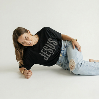 Woman lying on side laughing, wearing black "Jesus Saves" Luke 19:10 oversized Christian graphic tee with JESUS SAVES bold text design, paired with ripped denim jeans