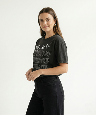 Woman standing in dark charcoal "Made to Worship" Christian graphic tee with bold WORSHIP text stacked design, paired with black wide-leg pants