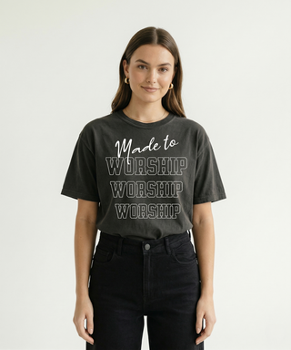 Front-facing view of woman in dark charcoal "Made to Worship" Christian graphic tee with stacked bold WORSHIP text design, paired with black jeans
