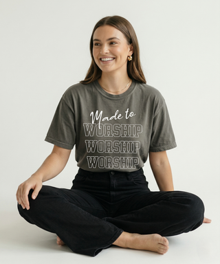 Smiling woman sitting cross-legged in charcoal "Made to Worship" Christian graphic tee with stacked WORSHIP bold text design, styled with black jeans