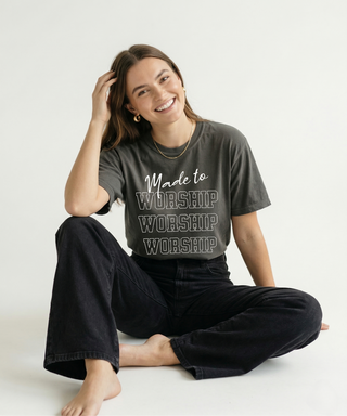 Woman sitting cross-legged hand in hair, smiling in charcoal "Made to Worship" women's Christian graphic tee with stacked WORSHIP text, black jeans