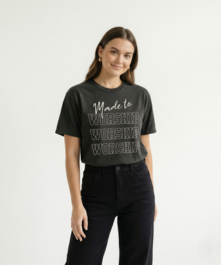 Woman standing smiling in dark charcoal oversized "Made to Worship" Christian graphic tee with stacked bold WORSHIP text, paired with high-waist black jeans