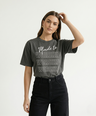 Woman with hand in hair in charcoal "Made to Worship" Christian graphic tee showing bold stacked WORSHIP design, styled with black jeans