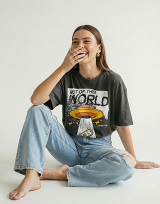 Laughing woman in charcoal "Not of This World" John 17:16 Christian graphic tee sitting casually with ripped denim jeans