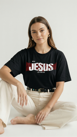 Woman sitting cross-legged in black "With Jesus Life Is Better" bold graphic Christian tee with red JESUS text design, styled with cream wide-leg trousers