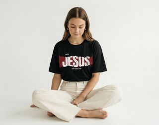 Close-up of woman sitting cross-legged showing "WITH JESUS LIFE IS BETTER" bold red Christian graphic tee design clearly, styled with cream wide-leg denim