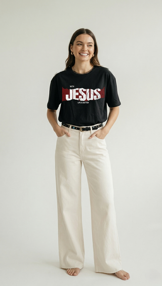 Full body view of smiling woman in black "With Jesus Life Is Better" oversized Christian graphic tee with bold JESUS print, paired with cream wide-leg trousers and belt