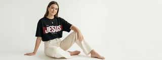 Woman sitting against white background in black "With Jesus Life Is Better" Christian graphic tee with JESUS bold red text, wearing cream wide-leg pants
