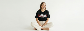 Smiling woman sitting cross-legged on white background in black "With Jesus Life Is Better" Christian graphic tee featuring bold red JESUS text print
