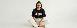 Woman sitting cross-legged with hands in lap wearing black "With Jesus Life Is Better" bold JESUS Christian graphic tee against clean white background